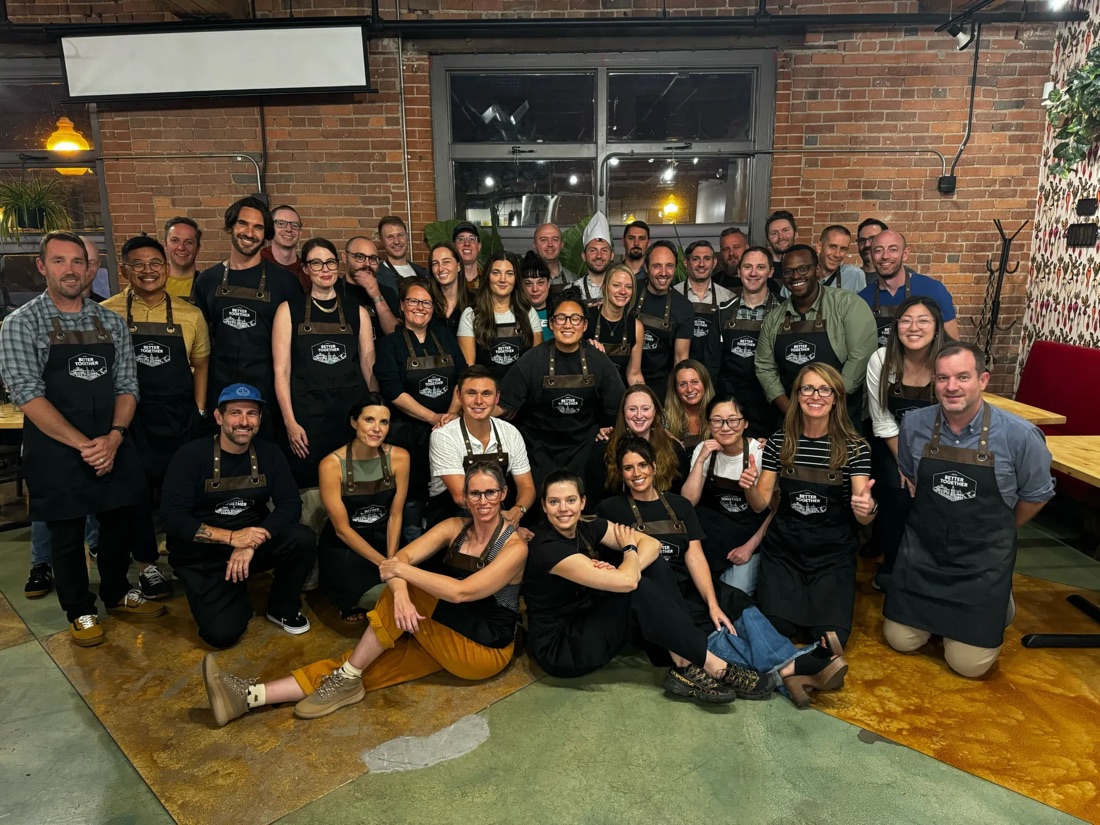 A large team in matching Better Together aprons gathered for a group photo in a brick-walled event space