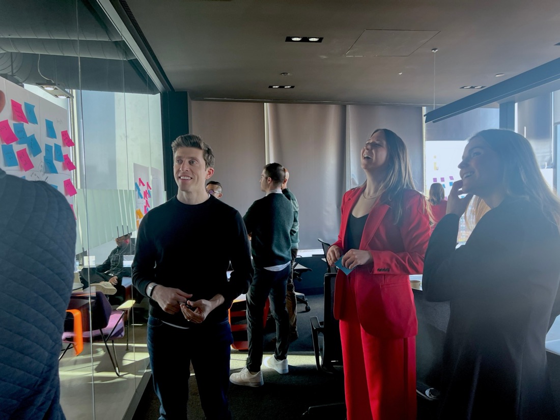 Colleagues laughing during a workshop in a glass-walled room covered in sticky notes