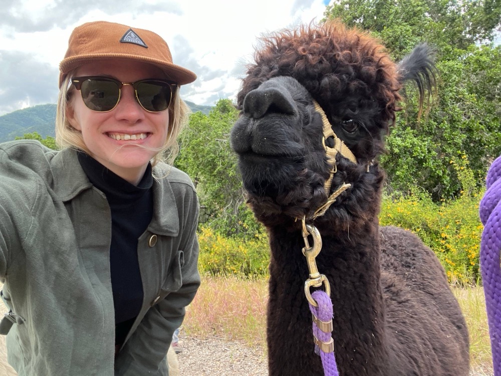 Beth in a cap and sunglasses, taking a selfie with a curly black alpaca