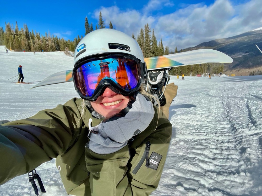 Beth grinning in a helmet and goggles on a ski slope, snowboard over her shoulder